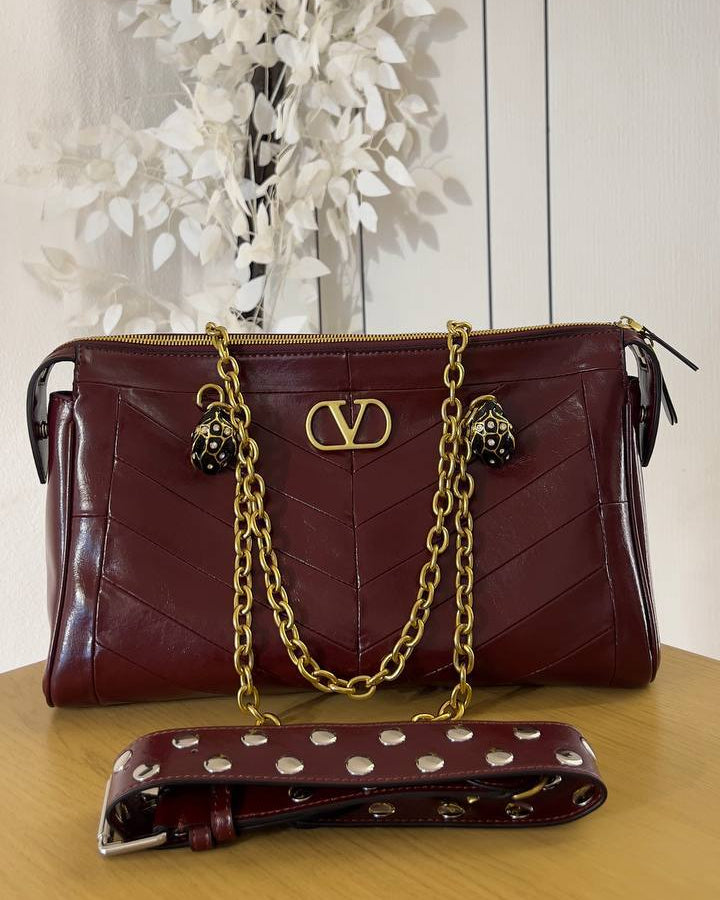 A burgundy quilted leather shoulder bag with gold-tone chain straps and a matching studded wristlet strap, displayed on a wooden surface against a white paneled background with decorative white leaves.
