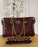 A burgundy quilted leather shoulder bag with gold-tone chain straps and a matching studded wristlet strap, displayed on a wooden surface against a white paneled background with decorative white leaves.
