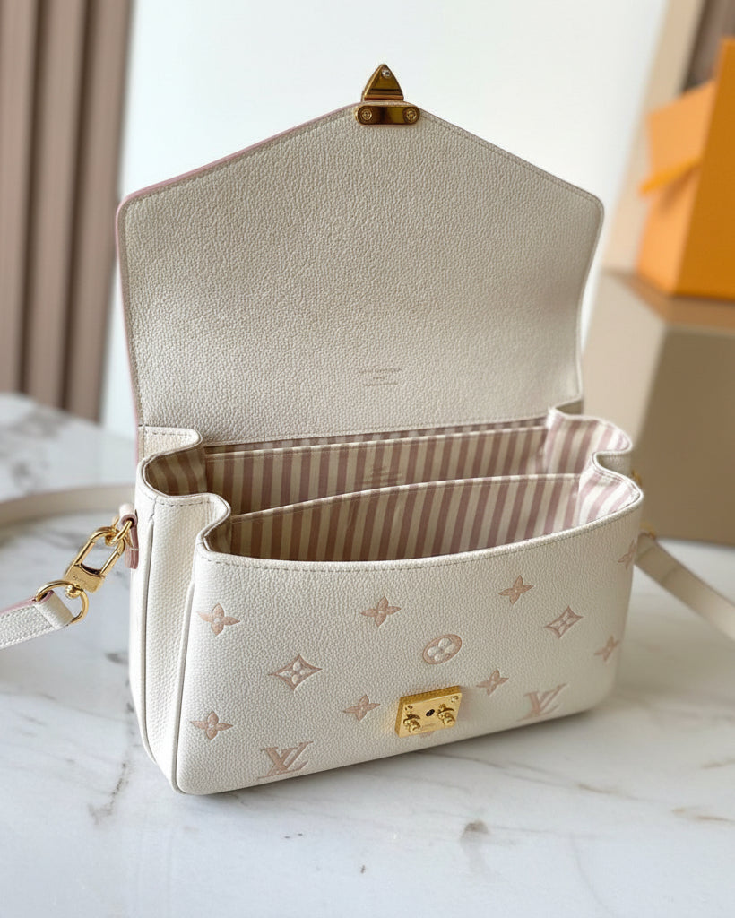 An open structured handbag in light beige pebbled leather with a subtle monogram pattern, gold-tone hardware, and a striped interior lining, photographed on a marble-like surface.