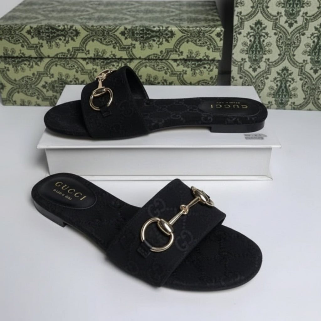 A pair of black Gucci flat slide sandals with the brand's signature GG monogram pattern and horsebit hardware, displayed on a white surface with a patterned green and white Gucci shoebox in the background.